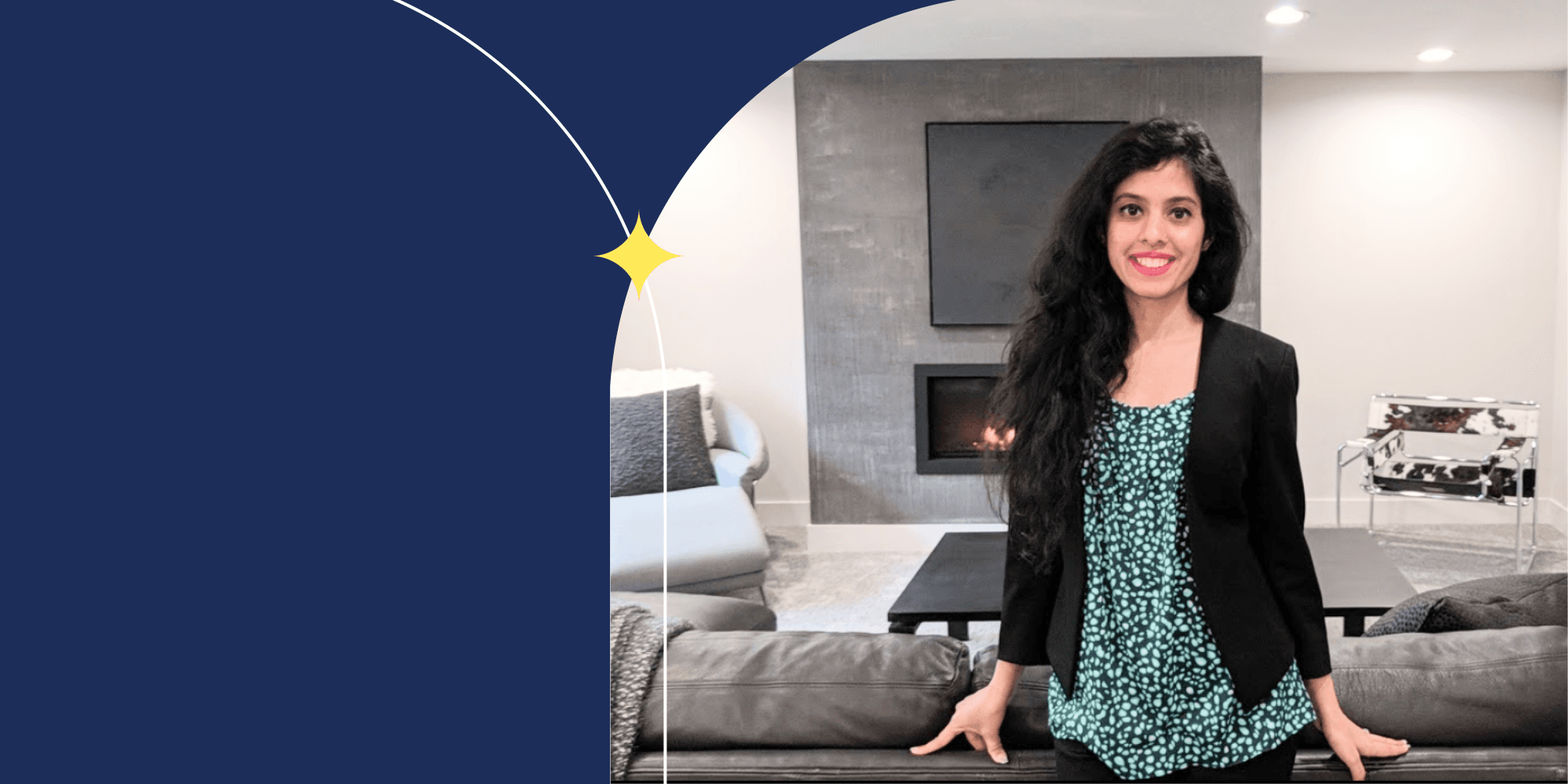 A smiling person standing in front of a couch. They wear a black blazer, a green blouse, dark pants, and pink lipstick. They have long hair. Behind them, there is a fireplace, a table, and two chairs.