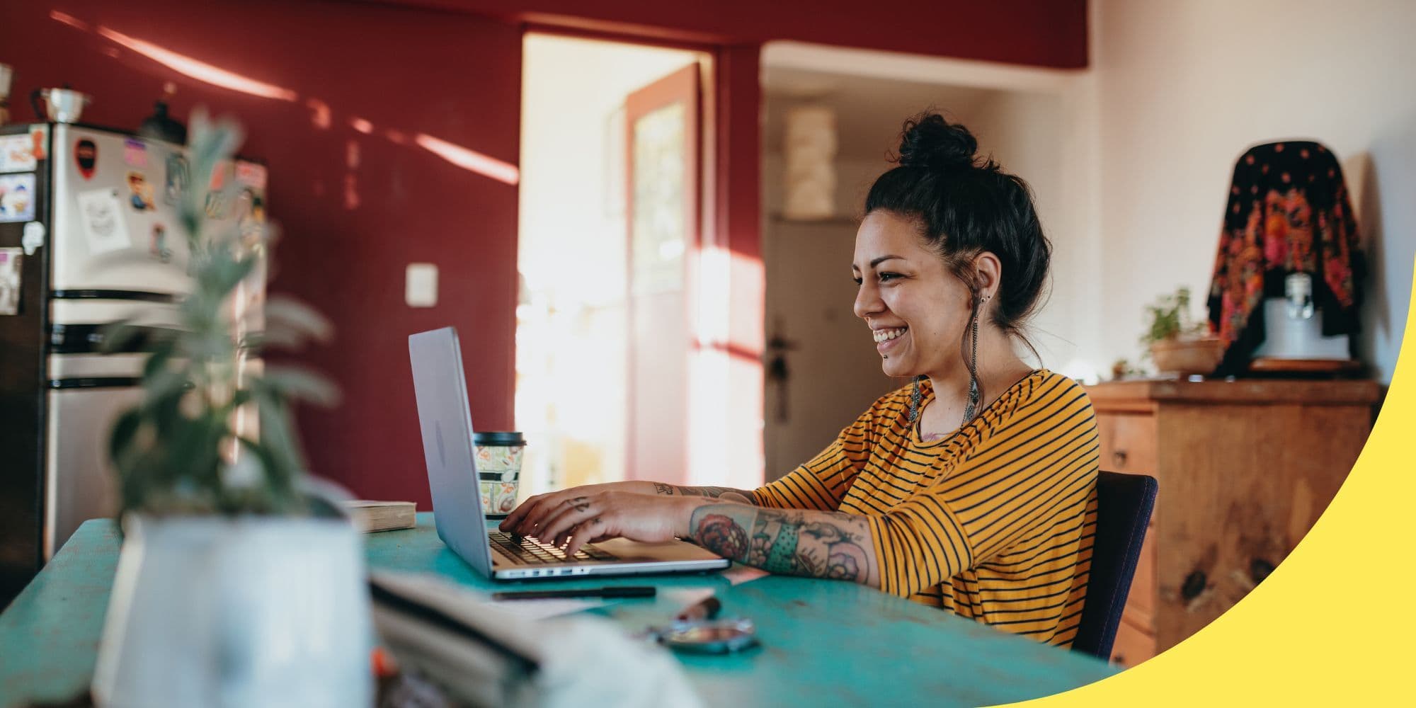 person sitting at a light blue table, typing on a laptop and smiling; red walls and a silver refrigerator are visible in the background