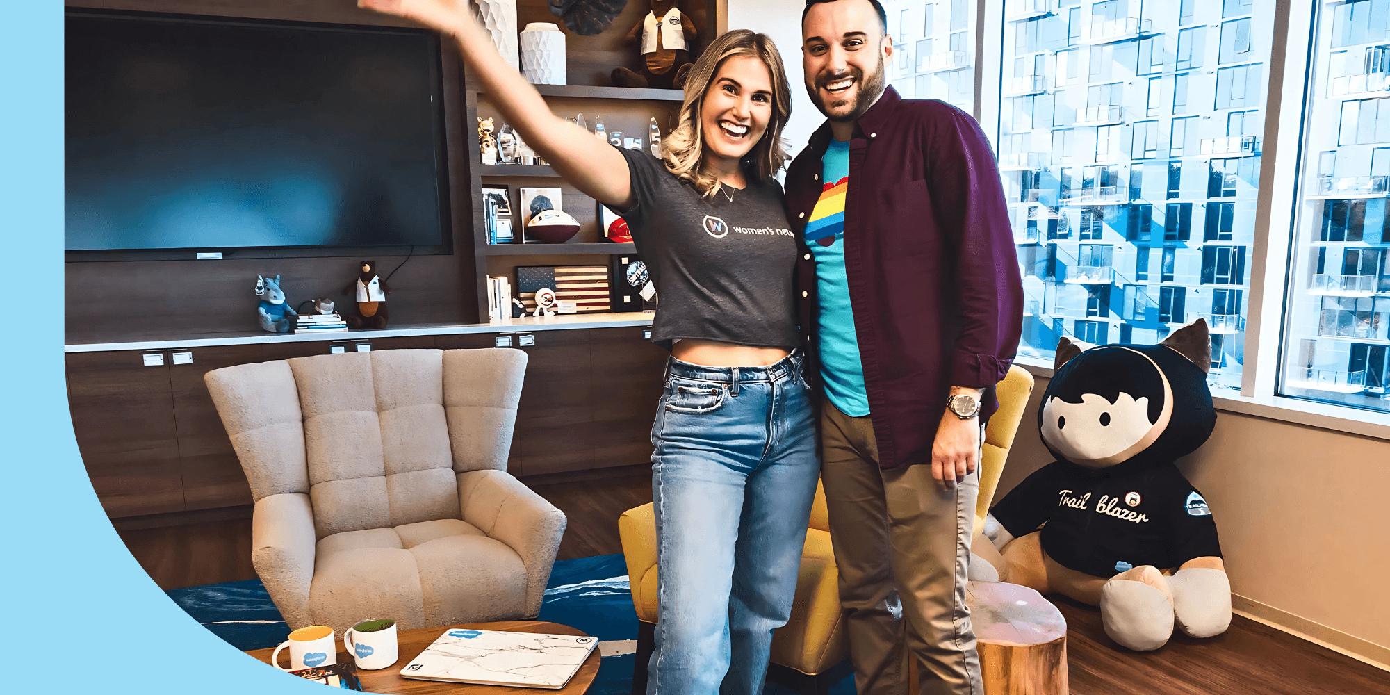 Two smiling people in an office. The person on the left wears a gray T-shirt, jeans, and white sneakers. They have shoulder-length hair. The person on the right wears a purple button-down shirt, a blue T-shirt, khakis, and dress shoes. They have short hair. Two chairs are behind them. To the far right, a black-and-white stuffed animal rests against the wall.