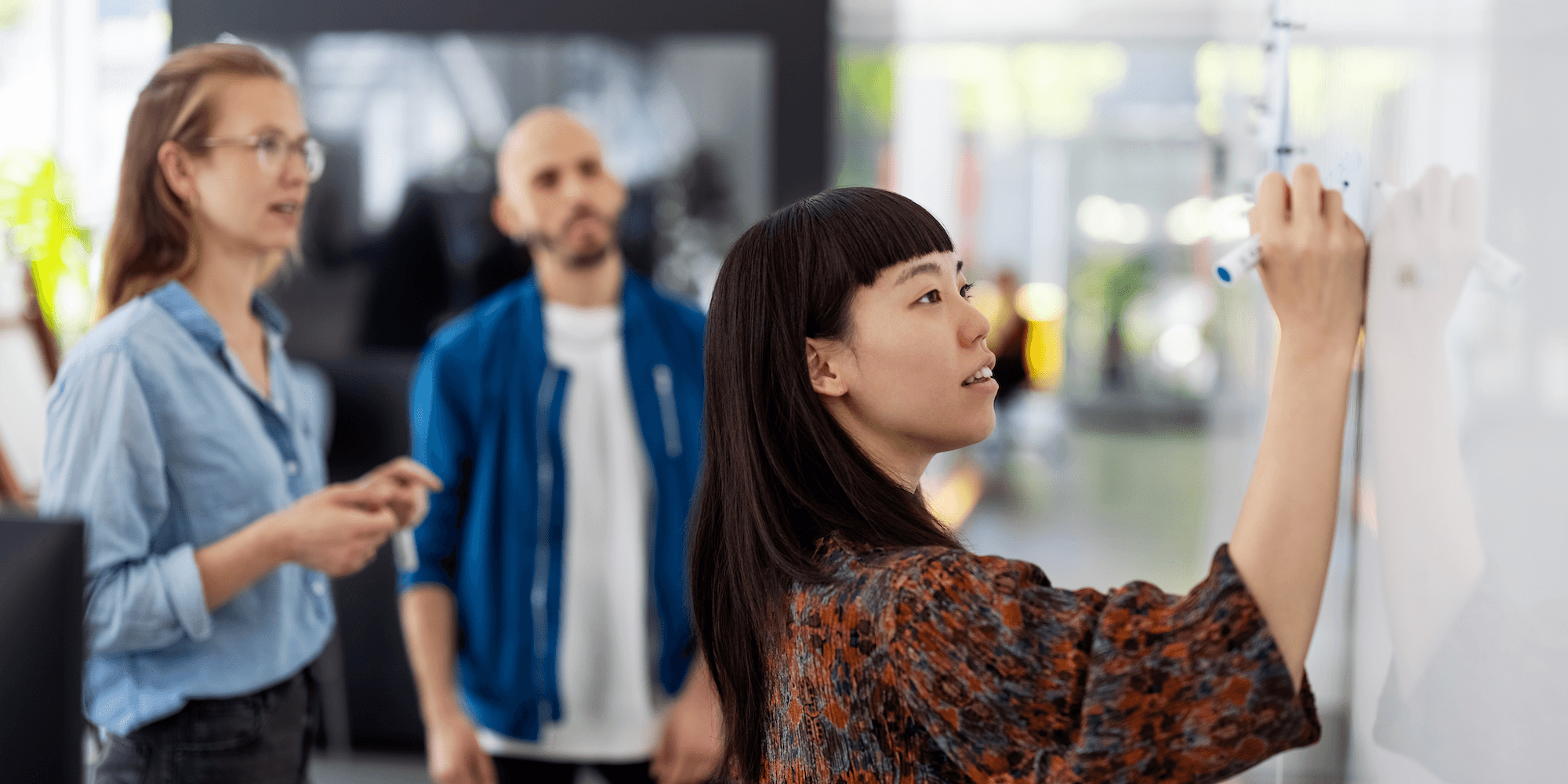 close-up of worker writing on a whiteboard during a meeting as two colleagues look on in the background